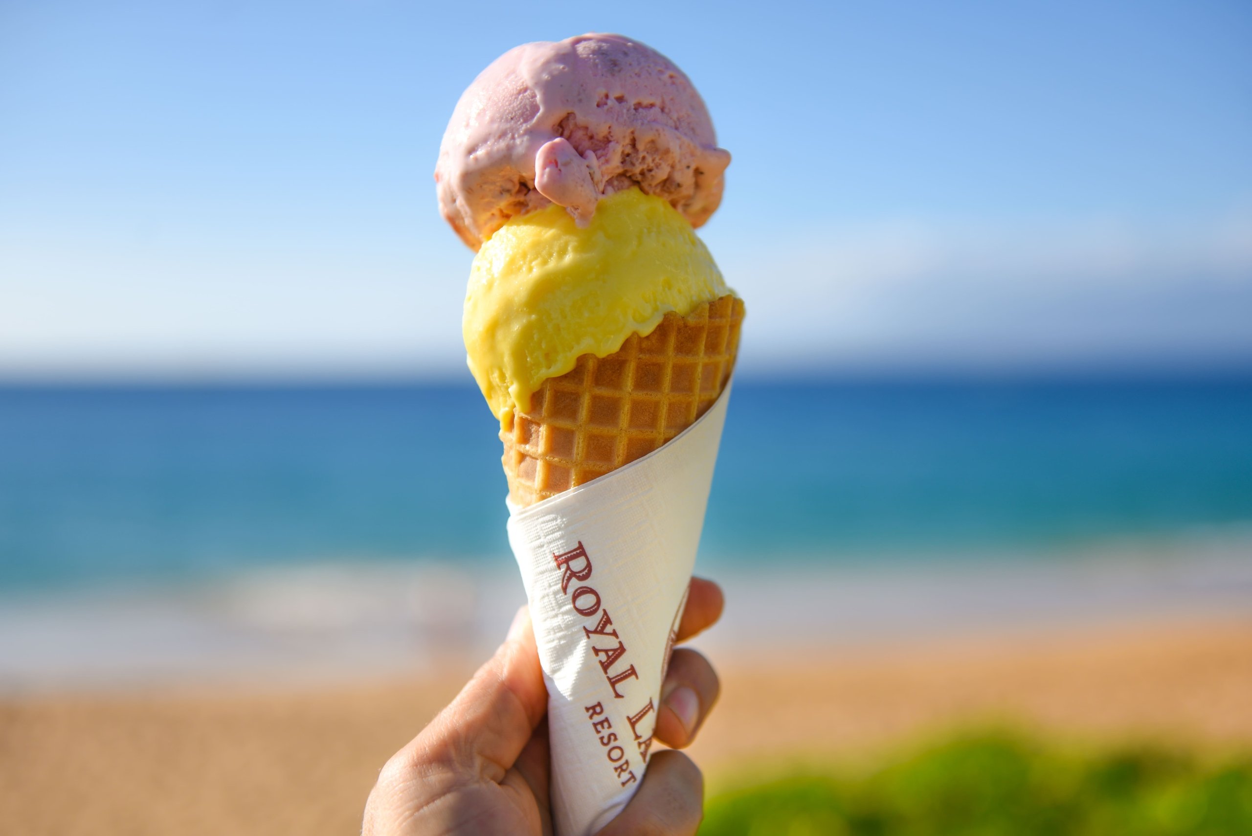 A person holding ice cream cone with two scoops and Maui's beach at the background in a sunny day