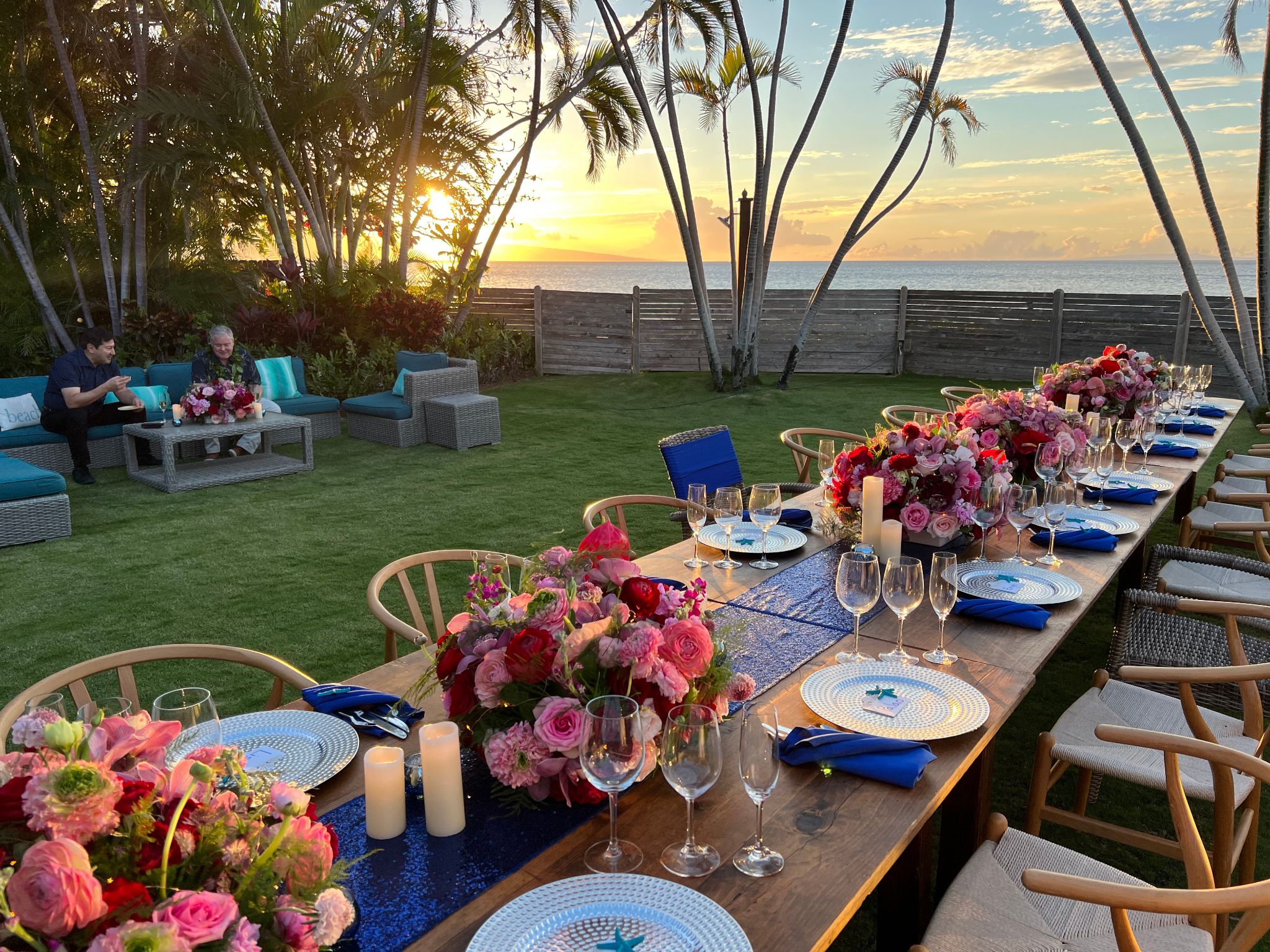 A beautifully set outdoor dining table with flowers, wine glasses, and candles, overlooking the ocean at sunset.