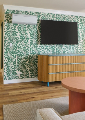 A wide view of a room with a green and white leaf-patterned wallpaper, a wooden dresser with a TV on top, and a wooden floor.