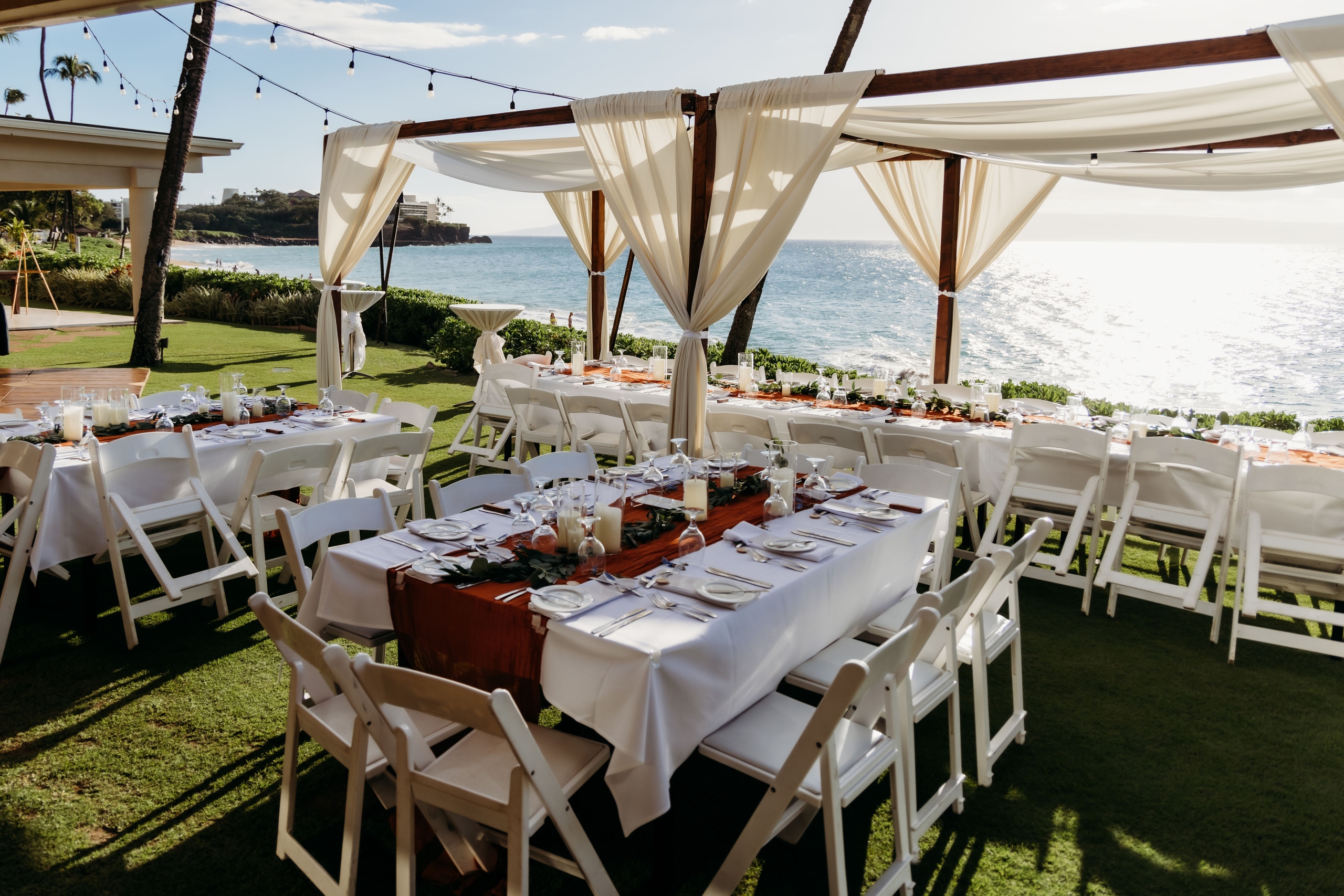 An outdoor dining area with white tables and chairs under a tent overlooking the ocean.