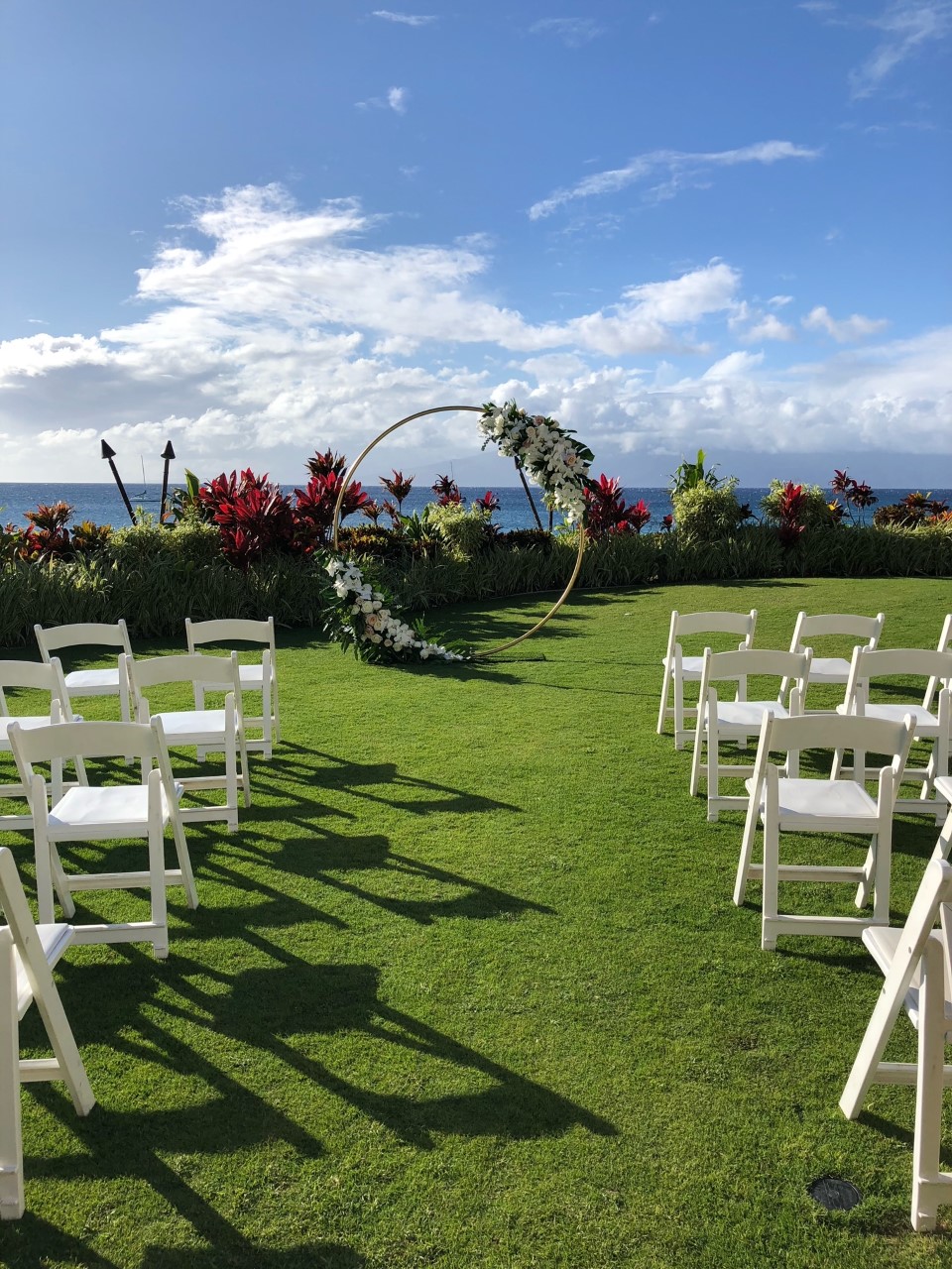 A wedding setup on a lawn with a floral arch, chairs, and an ocean view.