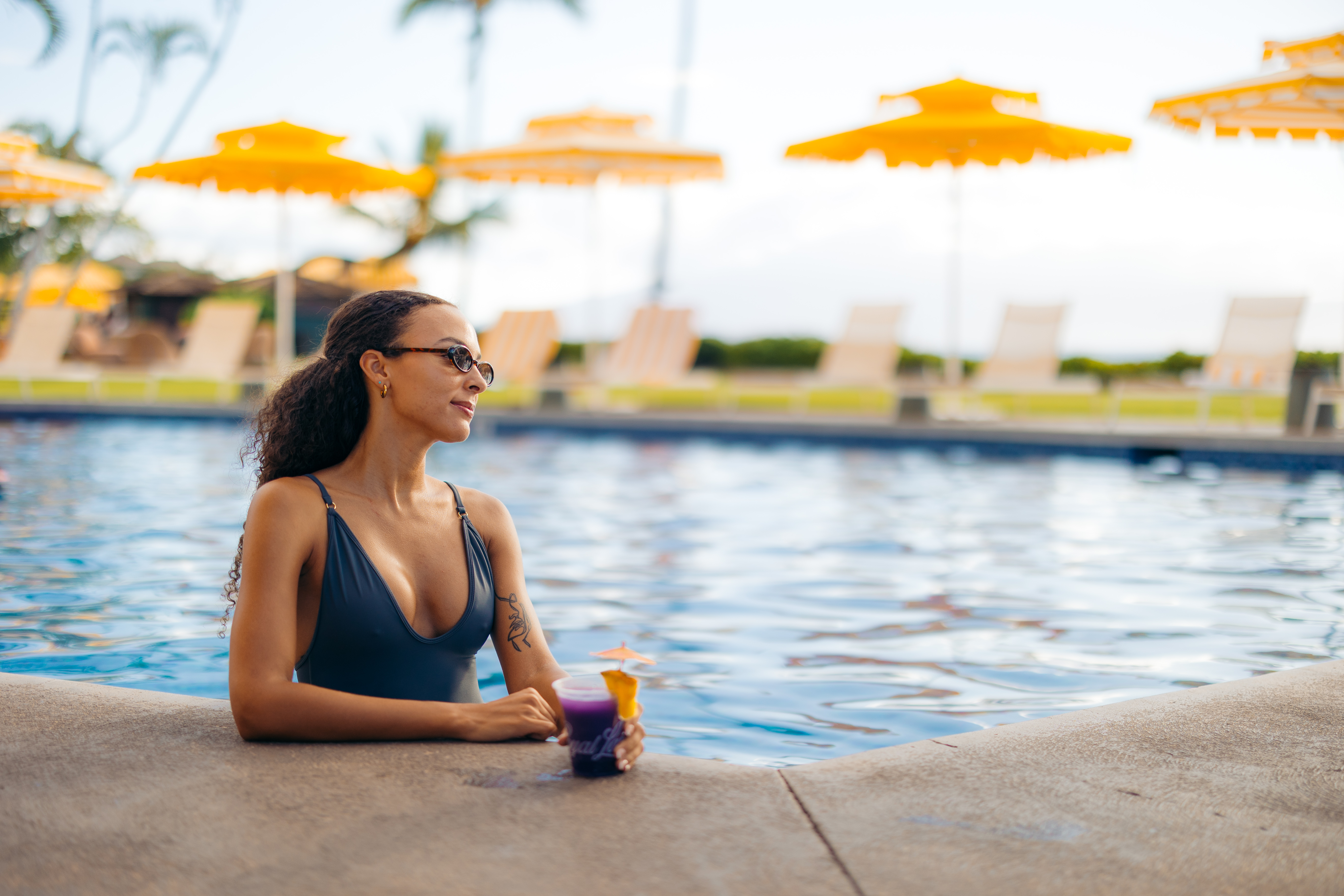 A woman in a swimsuit is sitting at the edge of a pool with yellow umbrellas and lounge chairs in the background.